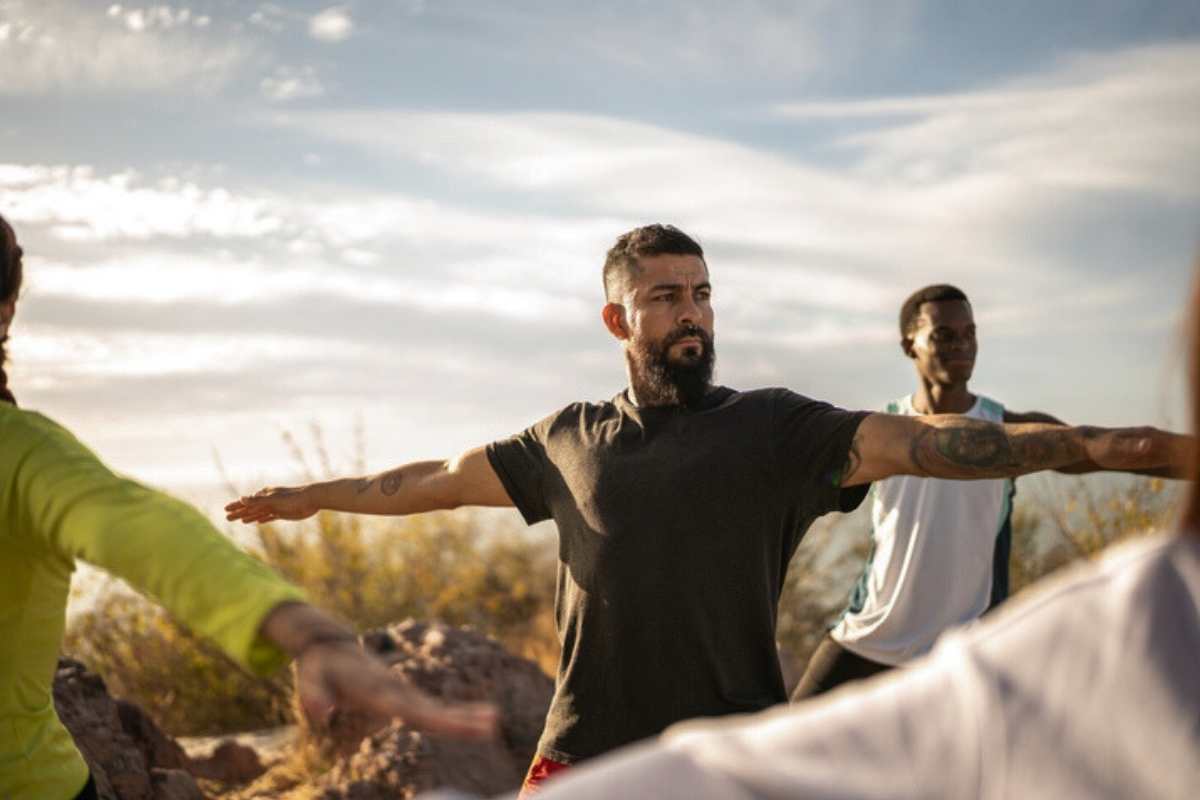 Veterans participate in an outdoor yoga session, standing with arms extended in a balanced pose beneath an open sky.