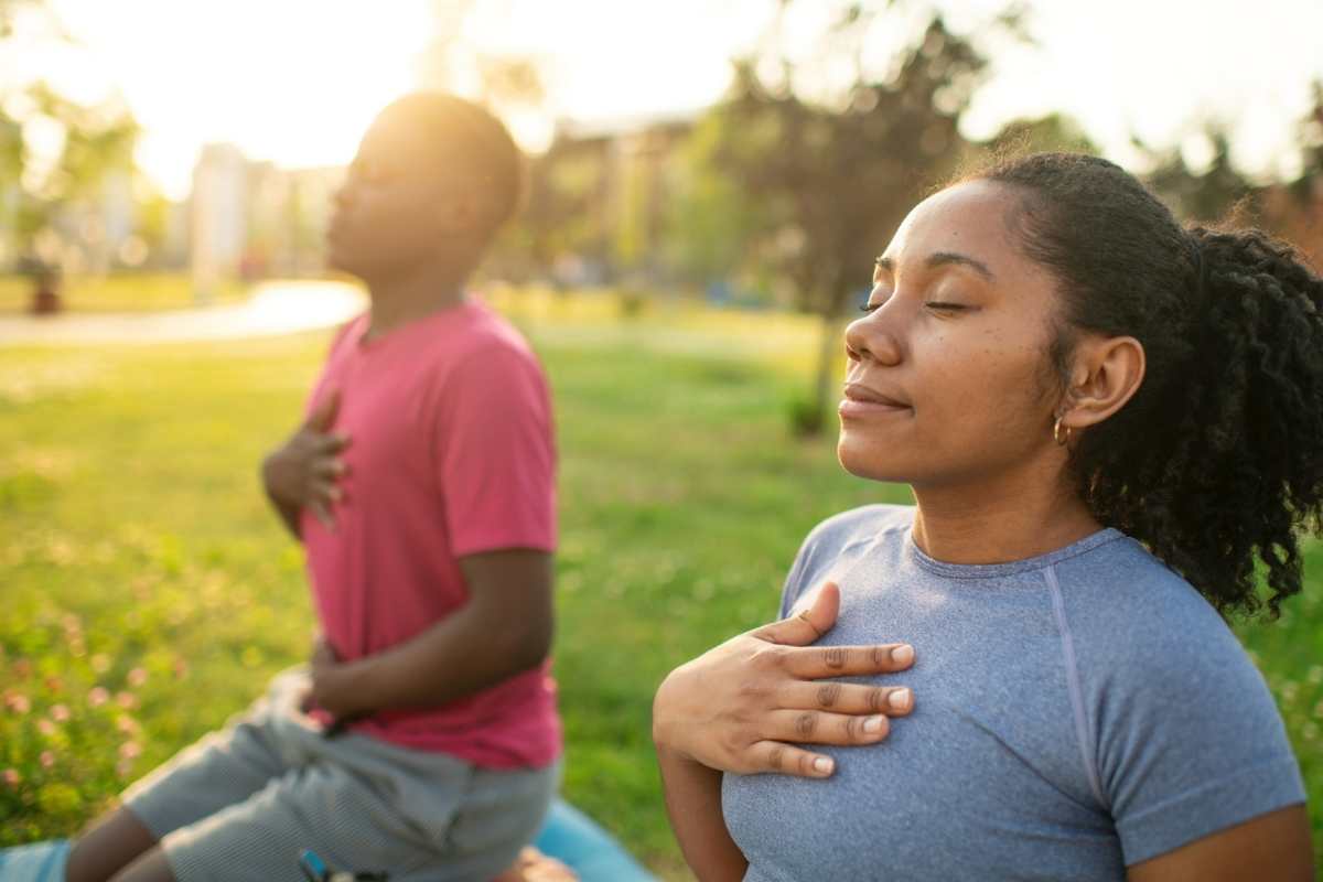 A woman veteran sits outdoors in a park, practicing mindful breathing with eyes closed and hands resting on their chests.