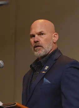 A bald man with a gray beard stands at a podium with a microphone, wearing a navy suit and looking serious while addressing an audience.