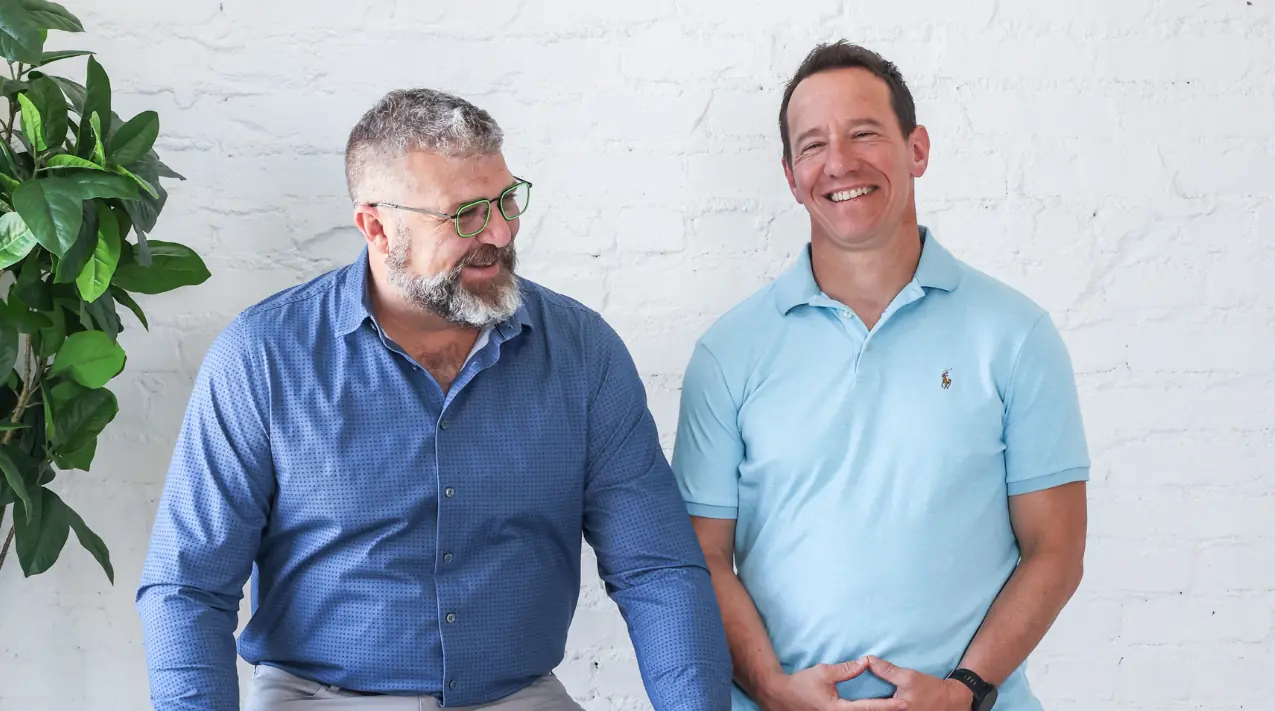 Two podcast hosts smiling and talking while standing against a white brick wall. One is wearing a blue button-down shirt and glasses, and the other is wearing a light blue polo shirt. A green leafy plant is visible on the left.