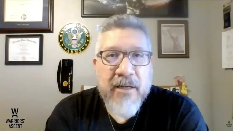 A man with glasses and a gray beard speaks on camera from an office, with military certificates and an Army emblem displayed on the wall behind him during a Warriors’ Ascent story recording.
