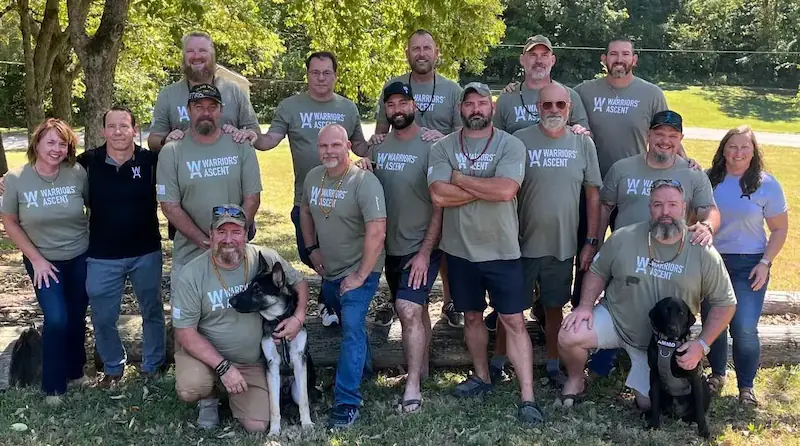 A photo of smiling men under a tree at a campground with sunshine and two dogs wearing branded Warriors' Ascent t-shirts